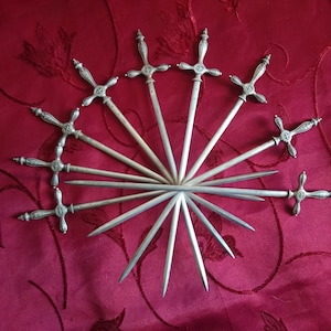 Peut inclure: Ensemble de huit épées à cocktail argentées avec des poignées ornées. Les épées sont disposées en éventail sur un fond de tissu rouge.