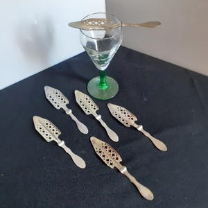 Peut inclure: Ensemble de barware vintage, comprenant un verre à absinthe transparent avec un pied vert et plusieurs cuillères à absinthe argentées. Les cuillères présentent des motifs ajourés décoratifs. Les articles sont disposés sur une surface sombre.