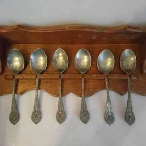 May include: A wooden wall-mounted shelf with six silver spoons hanging from it. The spoons have ornate designs and are arranged in a row.