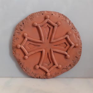Puede incluir: Una placa redonda de terracota con un diseño de cruz en relieve detallado. La cruz es de estilo tradicional con un círculo central y cuatro brazos ligeramente curvos.