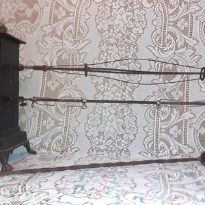 May include: An antique black metal rotisserie with a square base and a tall, rectangular body. It features a series of rods and wires for holding food, along with a long, slender poker. The background is a white lace tablecloth.