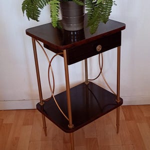Puede incluir: Una mesa auxiliar de madera oscura con un cajón y un estante inferior. La mesa tiene patas y detalles decorativos de metal dorado. Una planta de helecho en maceta encima. La mesa mide unos 61 cm de altura.