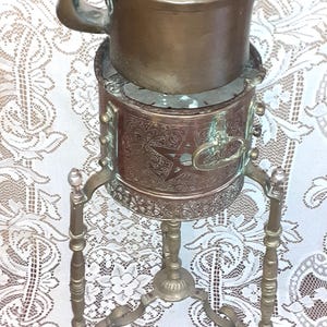 May include: A vintage copper tea kettle resting on a matching cylindrical copper stove stand. The stove features intricate engraved star and floral patterns, a side handle, and three slender, ornate metal legs with detailed finials.