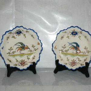 May include: Two white ceramic plates with blue scalloped edges. Each plate features a colourful bird design with green, yellow, blue, and orange accents. The plates are on black stands.