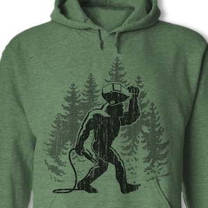 May include: Green hooded sweatshirt with a black silhouette of a Bigfoot holding a beer bottle in a forest setting.