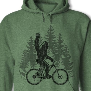 May include: Green hooded sweatshirt with a black silhouette of a Bigfoot riding a bicycle through a forest.