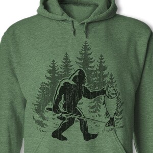 May include: Green hooded sweatshirt with a black silhouette of Bigfoot holding a fish in a forest setting.