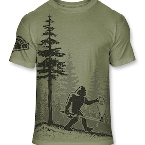 Puede incluir: Camiseta verde oliva con un gráfico negro de Bigfoot sosteniendo un pez y una caña de pescar en un bosque. El diseño incluye un árbol grande y árboles más pequeños. Una huella de tortuga está en la manga izquierda.
