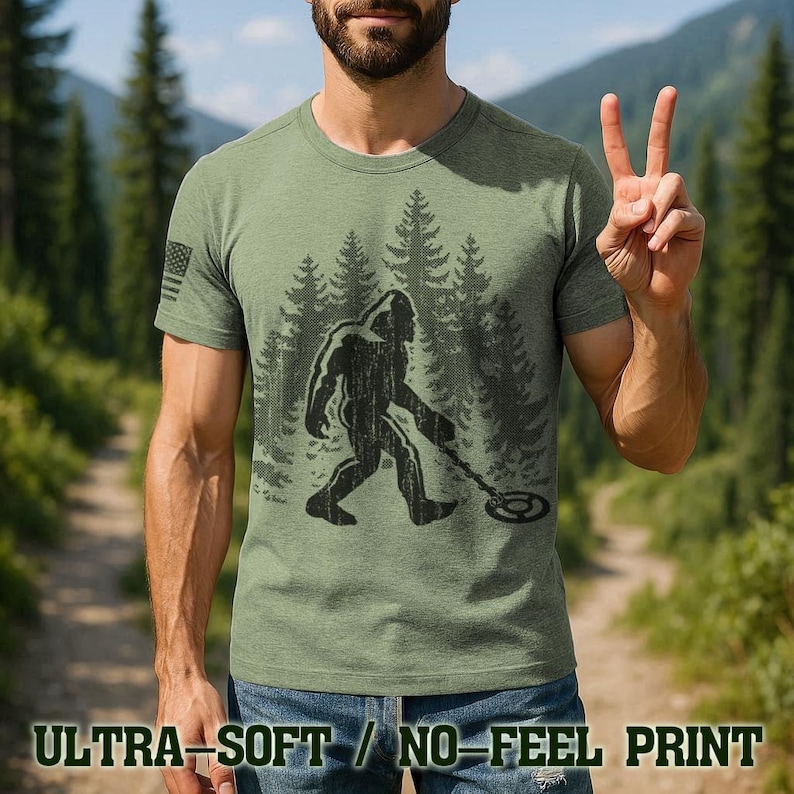 A man wearing a green t-shirt with a graphic of a Sasquatch figure and the text "Ultra-Soft / No-Feel Print" is making a peace sign with his hands.