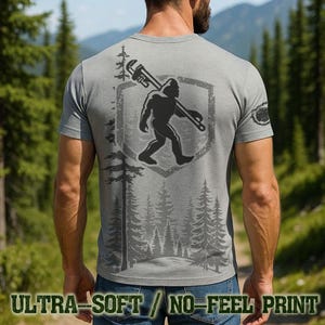 May include: Gray t-shirt featuring a Bigfoot graphic carrying a wrench, set within a shield design. The shirt includes the text "ULTRA-SOFT / NO-FEEL PRINT" at the bottom. The design incorporates forest imagery, creating a rugged, outdoorsy aesthetic.