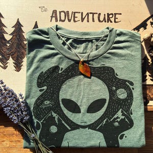 May include: Sage green t-shirt with a black graphic of an alien head, mountains, and planets. A leaf pendant hangs from the neckline. The wooden surface behind the shirt has the words "To Adventure" burned into it.