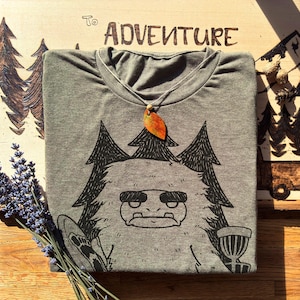 May include: A gray t-shirt with a black graphic of a cartoon yeti holding a frisbee and a disc golf basket. A leaf pendant hangs from the shirt's neckline. The word "ADVENTURE" is visible on the wooden surface.
