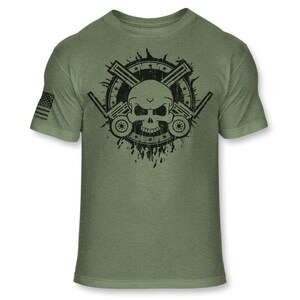 May include: Olive green t-shirt featuring a black graphic of a skull with crossed machine guns and a star-studded circle. The left sleeve displays a small American flag. The shirt is made of a soft, comfortable material.