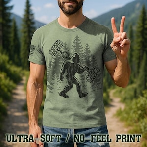 A man wearing a green t-shirt with a graphic of a Sasquatch and tires on it, making a peace sign with his hands.