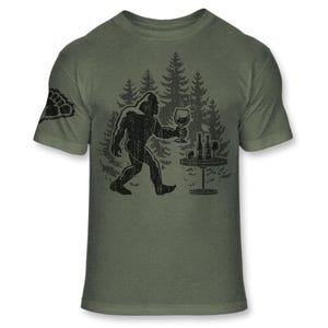 May include: Olive green t-shirt featuring a black graphic of Bigfoot holding a wine glass, walking towards a table with wine bottles and glasses. A forest silhouette is in the background. The left sleeve has a paw print.