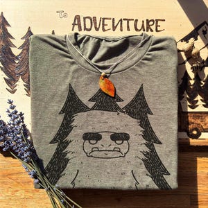 May include: Gray t-shirt with a black graphic of a cartoon yeti with pine trees. A leaf pendant hangs from the neckline. The word "ADVENTURE" is etched on a wooden surface in the background.