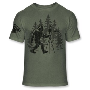 May include: Olive green t-shirt featuring a black graphic of Bigfoot holding a camera on a tripod in front of a forest scene. The left sleeve has a paw print design. A humorous design for nature lovers and photography enthusiasts.