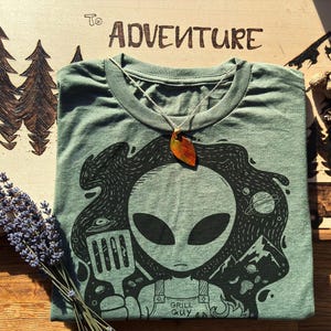 May include: Sage green t-shirt with a black graphic of an alien grilling. The alien is holding a spatula and a slice of pizza. A leaf pendant hangs from the shirt. The text "To Adventure" is visible on the wooden surface.