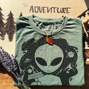 May include: A sage green t-shirt featuring a black graphic of an alien with space and mountain elements. A leaf pendant hangs on a string. The wooden surface behind the shirt has the words "To Adventure" and a mountain design.