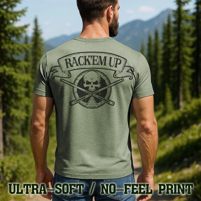 A man wearing a green t-shirt with a graphic design on the back, standing in a forested area with mountains in the background.