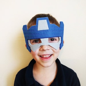 K&ouml;nnte beinhalten: Ein Kind tr&auml;gt eine blaue und graue Filzmaske mit einem Roboterdesign. Die Maske bedeckt die Augen des Kindes und einen Teil seiner Stirn.