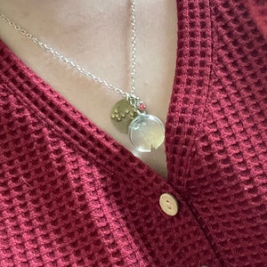 Op de afbeelding: Een zilveren ketting met een ronde glazen hanger met een gedroogde paardenbloem en een kleine ronde messing bedel met een stippenpatroon. De ketting wordt gedragen door iemand die een kastanjebruine wafel gebreide trui draagt.