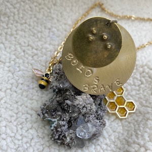 May include: Gold necklace with a round pendant that says "Bold & Brave". The pendant is layered with a smaller round pendant with a bee charm attached. The necklace is resting on a cluster of white crystals and a gold honeycomb charm.