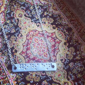 May include: A silver chain necklace with a silver bar pendant featuring Braille dots. The necklace is on a patterned rug.