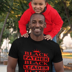 Puede incluir: Una camiseta negra con una corona roja y las palabras "Black Father Black Leader Black King" impresas en la parte delantera. Una ni&ntilde;a est&aacute; sentada sobre los hombros del hombre.