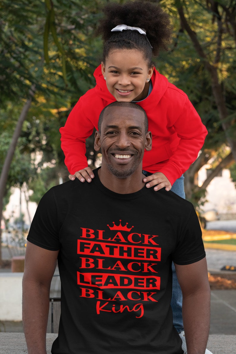 Puede incluir: Una camiseta negra con una corona roja y las palabras "Black Father Black Leader Black King" impresas en la parte delantera. Una ni&ntilde;a est&aacute; sentada sobre los hombros del hombre.