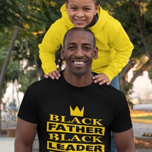 Puede incluir: Una camiseta negra con una corona dorada y las palabras "Black Father Black Leader Black King" impresas en la parte delantera.
