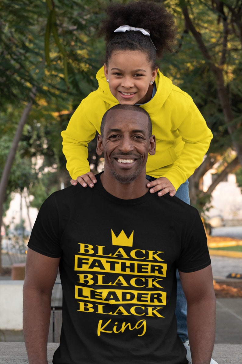 Puede incluir: Una camiseta negra con una corona dorada y las palabras "Black Father Black Leader Black King" impresas en la parte delantera.