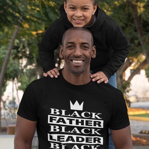 Puede incluir: Una camiseta negra con una corona blanca y el texto "Black Father Black Leader Black King" impreso en la parte delantera. Una ni&ntilde;a est&aacute; sentada sobre los hombros de un hombre que lleva la camiseta.