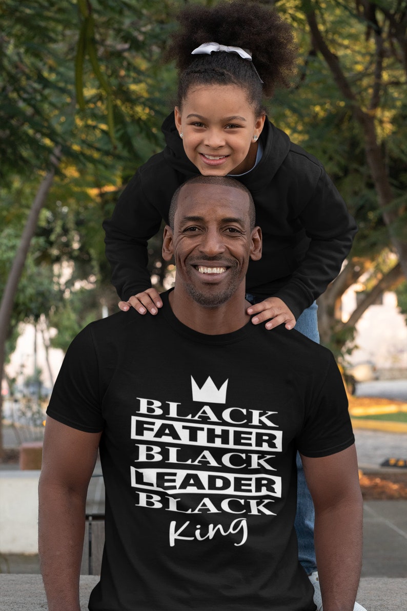 Puede incluir: Una camiseta negra con una corona blanca y el texto "Black Father Black Leader Black King" impreso en la parte delantera. Una ni&ntilde;a est&aacute; sentada sobre los hombros de un hombre que lleva la camiseta.