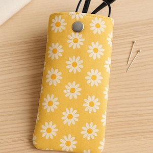 Puede incluir: Estuche amarillo para gafas con estampado de margaritas. El estuche de tela tiene un cierre de botón a presión. Las gafas negras son parcialmente visibles en el interior. Dos alfileres plateados están sobre la superficie de madera.