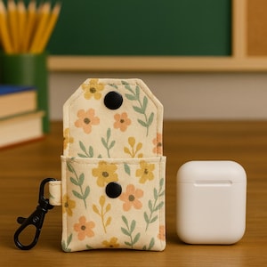 Könnte beinhalten: Beige AirPod-Hülle mit floralem Muster, schwarzem Verschluss und zwei schwarzen Druckknöpfen. Die Hülle ist geöffnet und gibt den Blick ins Innere frei. Eine weiße AirPod-Hülle liegt daneben auf einer Holzoberfläche.