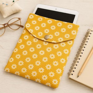 May include: Yellow tablet sleeve with a white daisy pattern, secured with a button closure. A tablet is partially visible inside. Other items include a pair of glasses, a notepad, and a pin cushion.