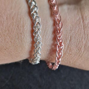 Peut inclure: Deux bracelets, l'un en argent et l'autre en cuivre, sont tissés en un motif tressé. Les bracelets sont sur le poignet d'une personne.