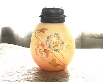 May include: A round, cream-coloured glass shaker featuring a hand-painted floral design of blue and pink pansies. The container is topped with a dark, perforated metal lid, providing a functional closure for dispensing contents from the glass vessel.