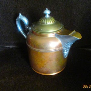 May include: A copper-colored teapot with a silver handle and spout. The lid is gold-colored with a silver finial. The spout has an ornate design. The teapot is likely vintage and would be a decorative item for a kitchen or dining room.