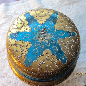 Op de afbeelding: Een ronde, vintage sieradendoos met een levendig ontwerp. De deksel heeft een turquoise midden met een bloemmotief, omgeven door gouden filigraan en accenten. De doos is waarschijnlijk van metaal.