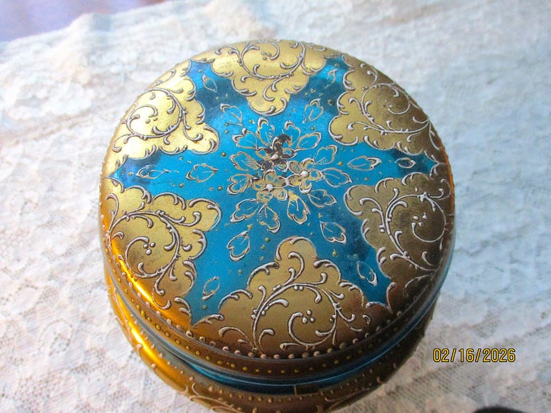 Op de afbeelding: Een ronde, vintage sieradendoos met een levendig ontwerp. De deksel heeft een turquoise midden met een bloemmotief, omgeven door gouden filigraan en accenten. De doos is waarschijnlijk van metaal.