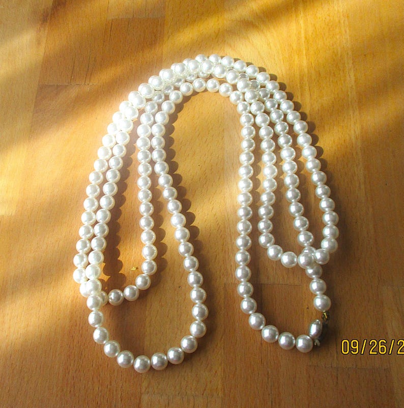 May include: A long, white pearl necklace with multiple strands, resting on a wooden surface. The pearls are round and evenly sized, with a slight sheen. The necklace is draped in a flowing pattern, catching the light.