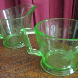 May include: A set of two vintage green glass creamer and sugar bowl with a floral design. The creamer has a handle and a spout. The sugar bowl has a handle and a rounded shape.
