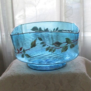 May include: A blue glass bowl with a wavy design and a painted rose vine. The bowl has a square shape and a wide base.