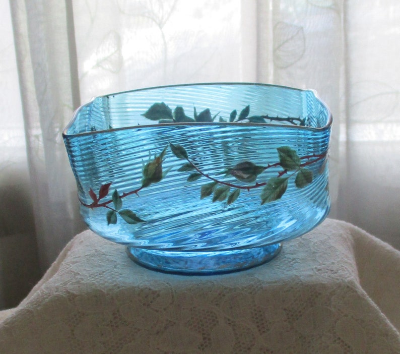 May include: A blue glass bowl with a wavy design and a painted rose vine. The bowl has a square shape and a wide base.