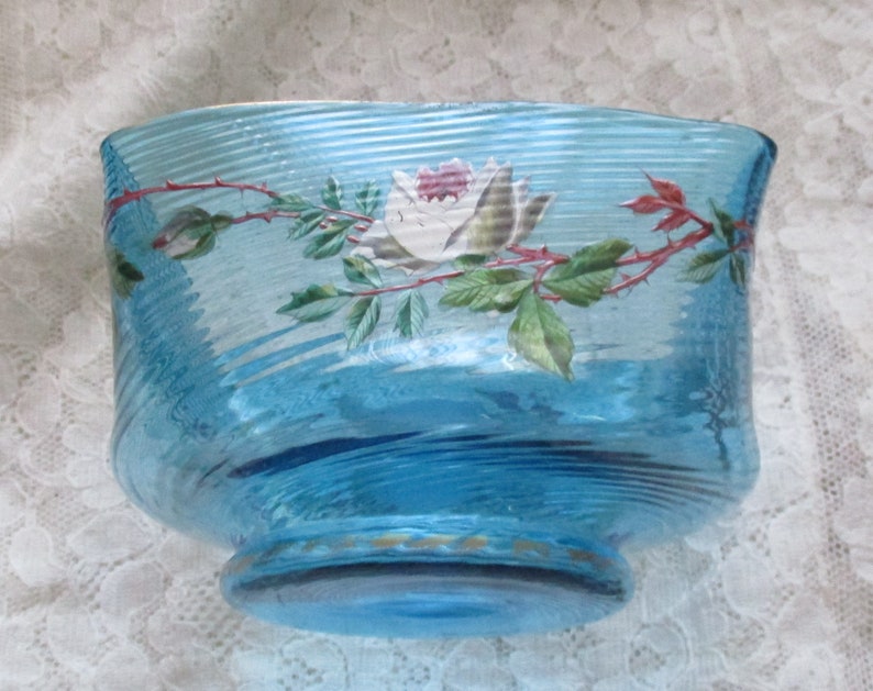 May include: A blue glass bowl with a white rose and green leaves painted on the side. The bowl has a wavy, textured surface.