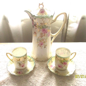 May include: A porcelain coffee pot with a lid and two matching cups and saucers. The set features a cream-colored base with pink rose floral patterns, green scalloped edges, and gold leaf accents. The date 07/21 is printed in the bottom right corner.