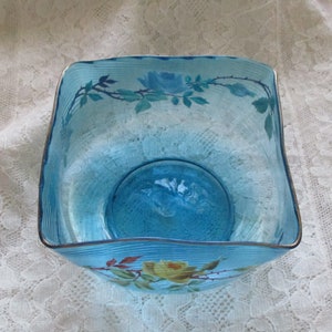 May include: A square, blue glass bowl with a wavy rim and painted floral designs. The bowl has a light blue tint and features delicate white and yellow roses with green leaves.