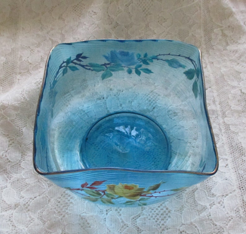 May include: A square, blue glass bowl with a wavy rim and painted floral designs. The bowl has a light blue tint and features delicate white and yellow roses with green leaves.